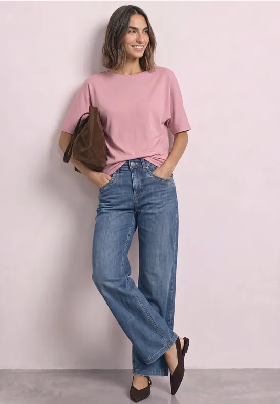 Vrouw in een roze T-shirt, blauwe jeans en zwarte schoenen, glimlachend met een bruine tas over de schouder.