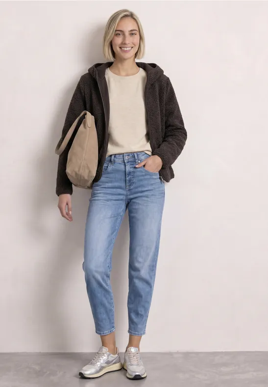 Frau in lässiger Kleidung: braune Jacke, weißes Shirt, Jeans, silberne Sneaker und Tasche vor heller Wand.