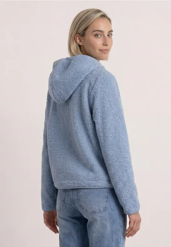 Frau von hinten im hellblauen Fleece-Hoodie und blauen Jeans, schaut über die Schulter.