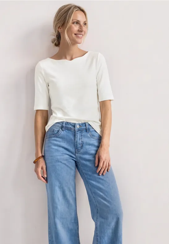 Une femme posant devant un fond clair, vêtue d'un haut blanc à manches mi-longues et d'un jean bleu large.