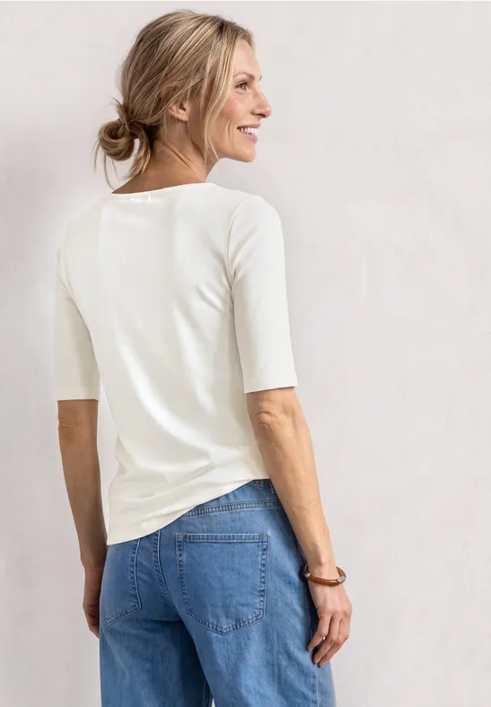 Une femme vue de dos porte un haut blanc à manches mi-longues et un jean bleu devant un mur clair.