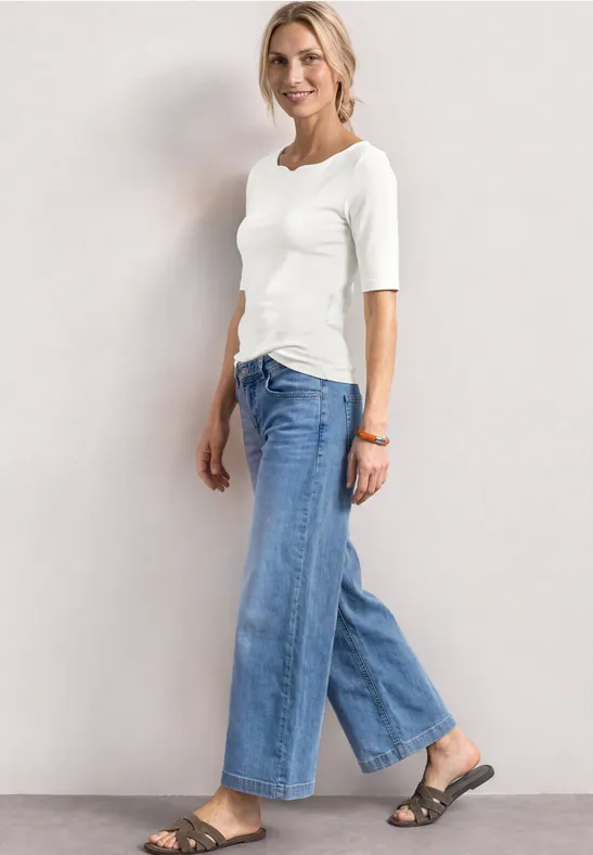 Femme en top blanc 3/4, jean large bleu et sandales, posant devant un fond neutre.