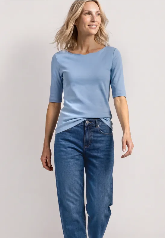 Een vrouw staat in een lichtblauw shirt en blauwe jeans voor een neutrale achtergrond.