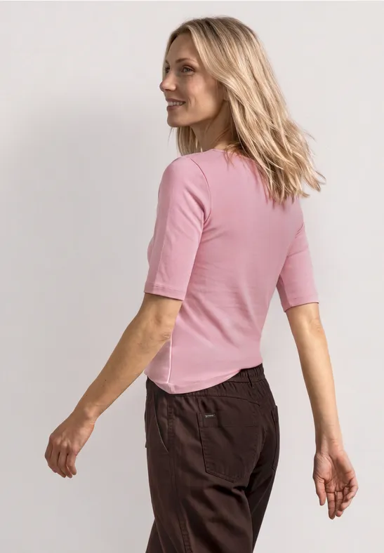Une femme de dos en haut rose à manches mi-longues et pantalon sombre marche de profil.