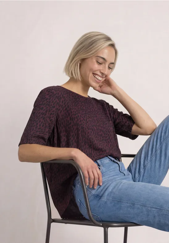 Jeune femme blonde souriante assise confortablement sur une chaise, portant un jean et un haut à motifs.