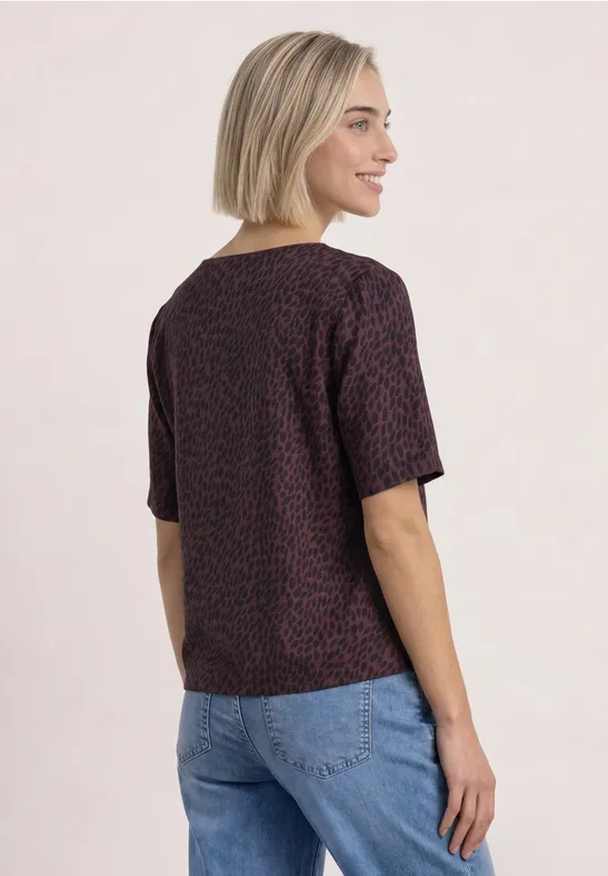 Femme aux cheveux blonds courts portant un t-shirt violet à petits motifs et un jean bleu, vue de profil.