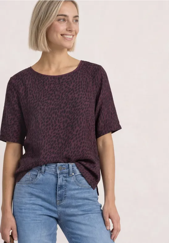 Femme en tee-shirt violet à motifs et jean, souriante devant un fond clair.