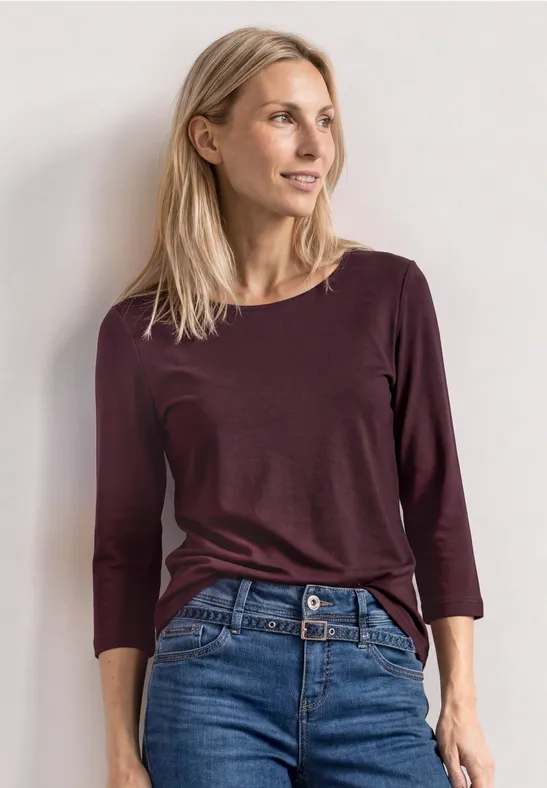 Eine schlanke Frau in burgunderfarbenem Langarmshirt und Jeans vor heller Wand.