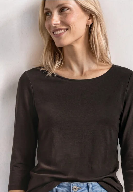 Eine lächelnde Frau in einem braunen Langarmshirt vor heller Wand.