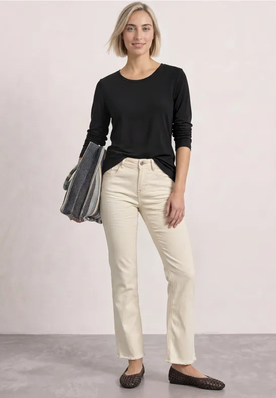 Eine lächelnde Frau in schwarzem Langarmshirt und beigen Jeans, mit Tasche in der Hand.