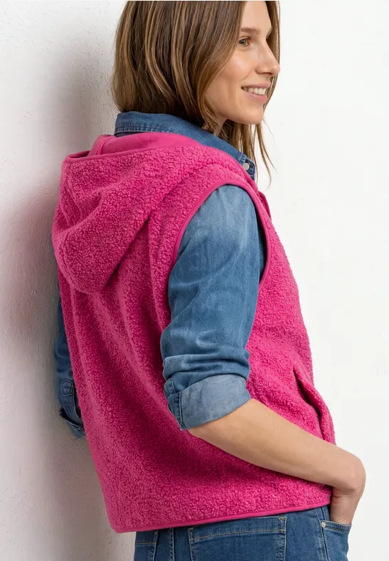 Frau in pinker, flauschiger Hoodie-Weste über Jeanshemd, seitlich stehend und lächelnd.