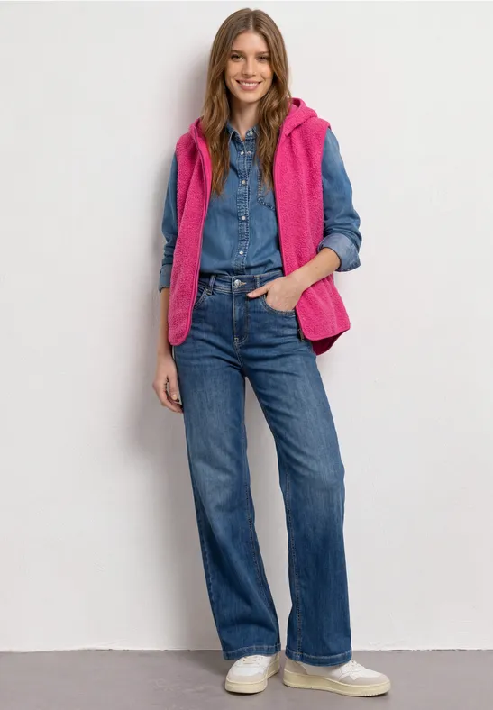 Frau in Jeanshemd und -hose, pinker Steppweste und weißen Sneakern, mit Händen in den Taschen.