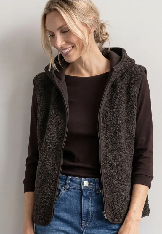 Lächelnde Frau in dunkelbraunem Fleece-Kapuzenweste über schwarzem Langarmshirt und Jeans.