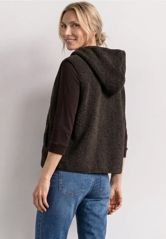 Lächelnde Frau mit blondem Haar in schwarzer Kapuzenweste, schwarzem Langarmshirt und blauer Jeans.