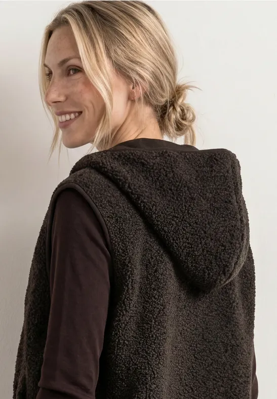 Eine lächelnde Frau mit blondem Zopf trägt eine flauschige braune Kapuzenweste über schwarzem Shirt.