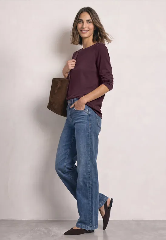 Frau in lässigem Look mit weiten Jeans, bordeauxfarbenem Pullover und brauner Tasche.