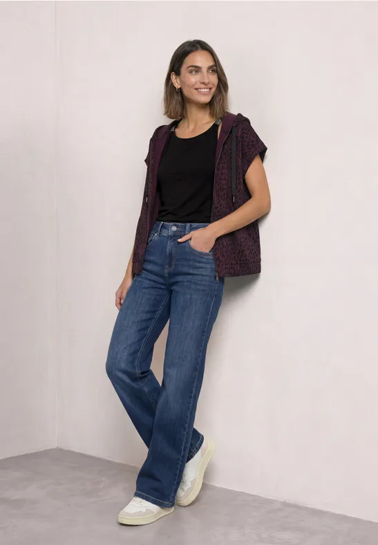 Frau in schwarzem T-Shirt mit karierter Weste, blauen Jeans und weißen Sneakern vor hellem Hintergrund.