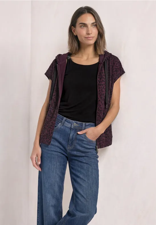 Frau steht lässig vor heller Wand, trägt schwarzes Top, violette Kapuzenweste mit Leopardenmuster und blaue Jeans.