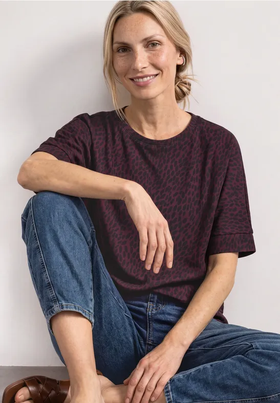 Eine lächelnde Frau sitzt entspannt in Jeans und einem gemusterten T-Shirt vor hellem Hintergrund.