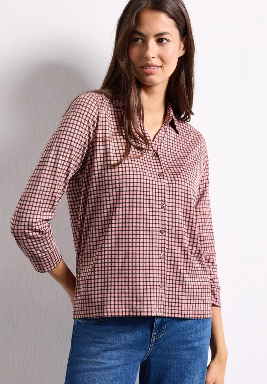 Une femme porte un chemisier à carreaux rouge et blanc et un jean bleu devant un mur blanc.