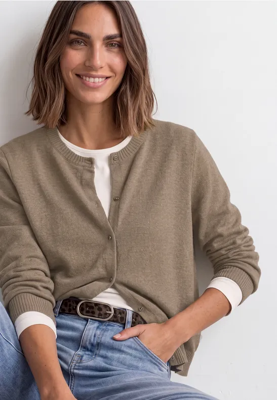 Modell sitzt lächelnd in Jeans, weißem Shirt und khaki Cardigan, akzentuiert mit Gürtel.