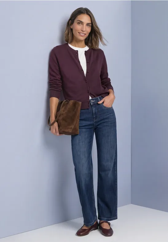 Frau lächelt, trägt weiße Bluse, violette Jacke, gerade Jeans, braune Loafers und hält eine braune Clutch.