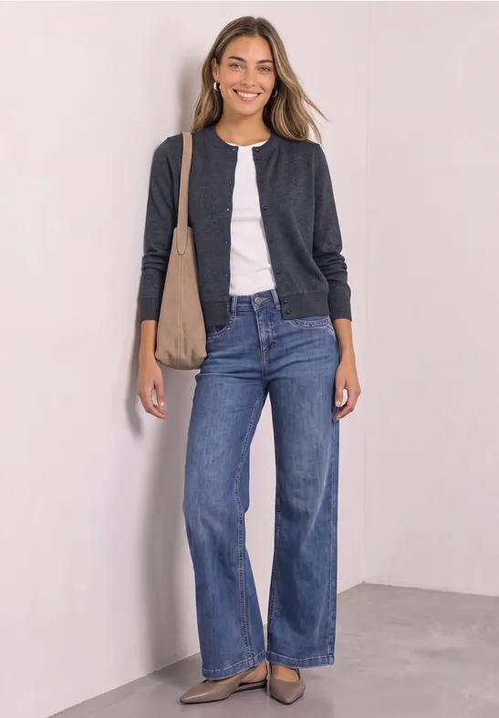 Vrouw lacht in jeans, wit shirt en donkergrijs vest met schoudertas tegen witte muur.