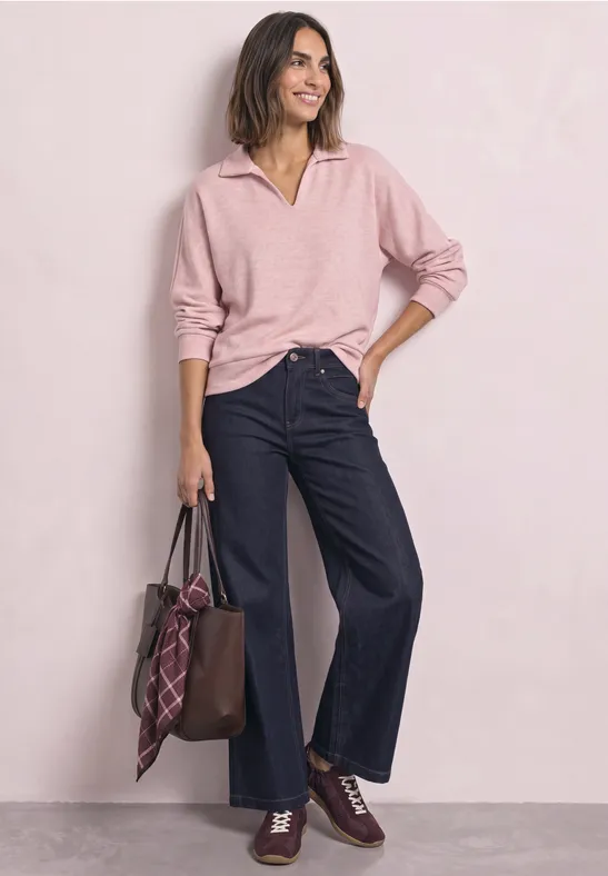Frau in rosa Pullover, weiten Jeans und Ledertasche posiert lächelnd vor hellem Hintergrund.