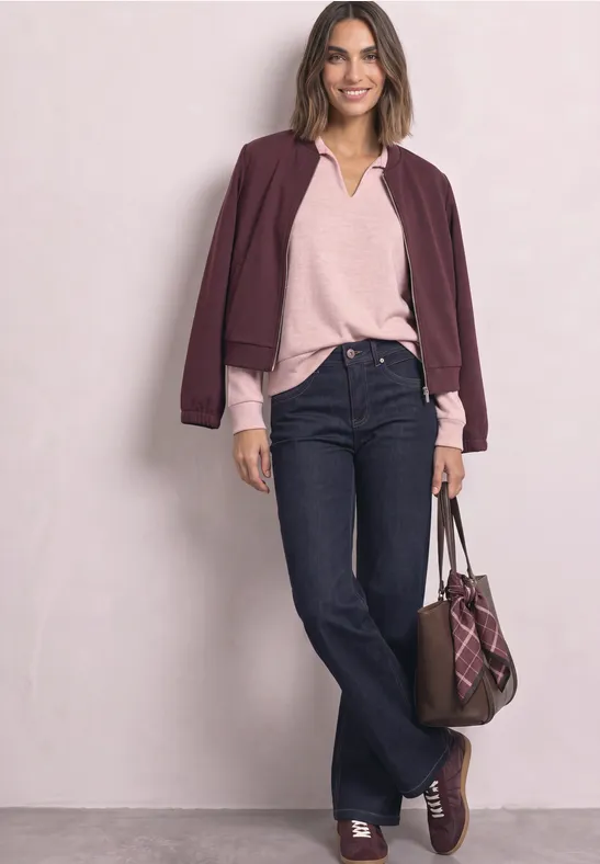 Frau in Jeans, rosa Shirt und bordeauxfarbener Jacke, mit passender Tasche und Schuhen, lächelnd.