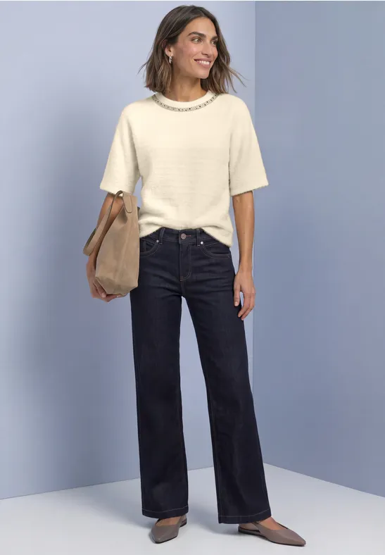 Frau mit cremefarbenem Kurzarmtop, dunklen weitem Jeans, braunen Schuhen und Tasche vor blauem Hintergrund
