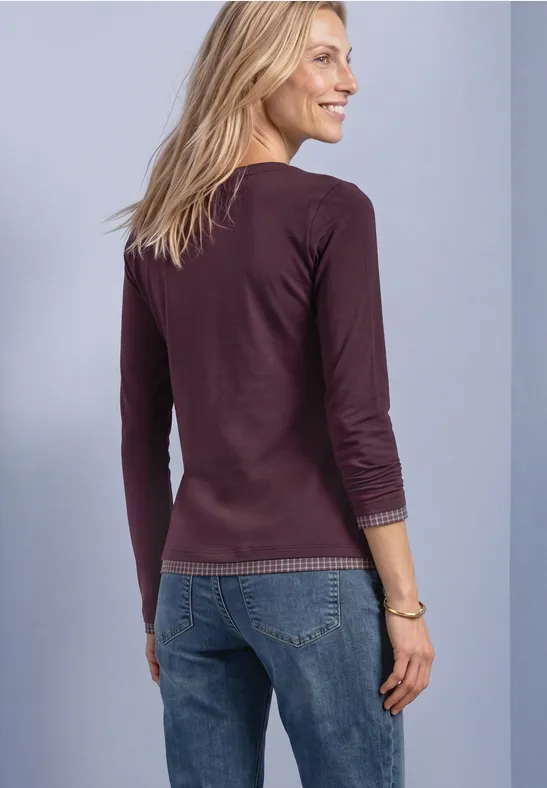 Frau von hinten halblinks in braunem Langarmshirt und blauen Jeans vor neutralem Hintergrund.
