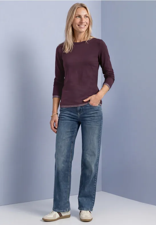 Eine lächelnde Frau in lila Pullover und blauen Jeans vor neutralem Hintergrund.