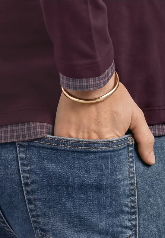 Ein Mensch steckt die Hand in die Gesäßtasche blauer Jeans, trägt burgunderrotes Oberteil und goldenen Armreif.