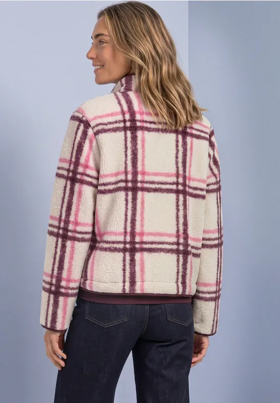 Frau trägt weiße, flauschige Jacke mit pink-lila Karomuster, steht seitlich vor hellblauem Hintergrund.