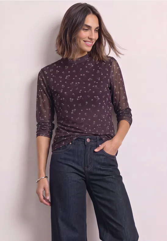 Frau in dunkelblauer, geblümter Spitzenbluse mit Mesh-Ärmeln und Jeans, lächelnd vor hellem Hintergrund.