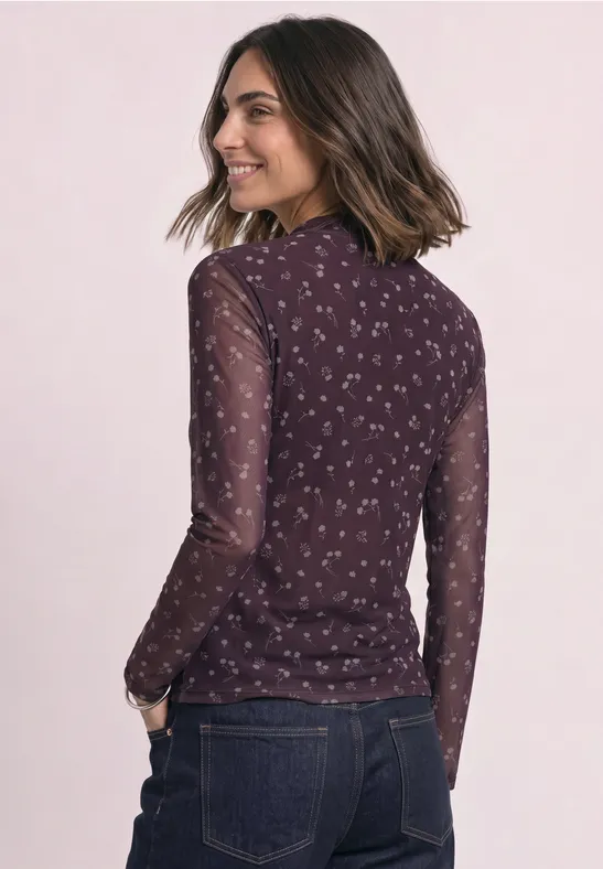 Eine Frau trägt eine dunkelviolette, halbtransparente Bluse mit Blumenmuster und Jeans, Rückenansicht.