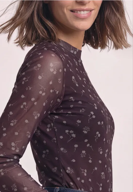 Nahaufnahme einer lächelnden Frau in langärmligem, leicht transparentem, dunklem Shirt mit kleinem Blumenmuster.