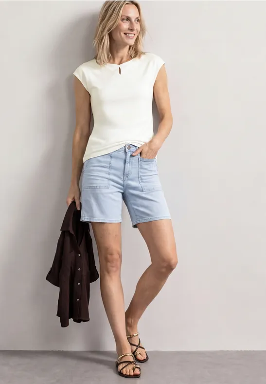 Frau in weißem Top und hellblauen Jeansshorts, hält eine braune Jacke, trägt schwarze Sandalen.