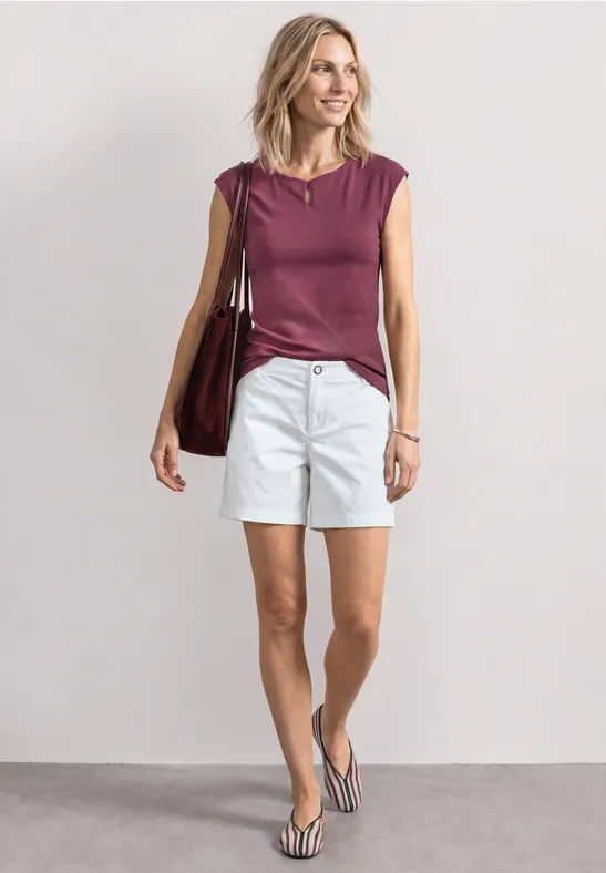 Femme en débardeur bordeaux, short blanc et tongs, portant un grand sac sur fond neutre.