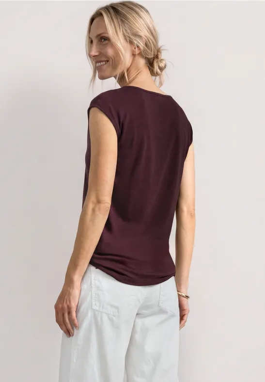 Femme de dos portant un top bordeaux sans manches et un pantalon blanc ample sur fond clair.