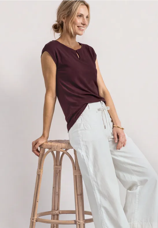 Femme appuyée sur un tabouret en bois, porte un haut bordeaux à manches courtes et un pantalon large clair.