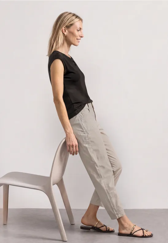 Femme en top noir et pantalon beige s’appuyant de côté sur une chaise, chaussures plates.