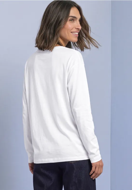 Frau mit schulterlangen Haaren trägt weißes Langarmshirt, steht seitlich und schaut über die Schulter.