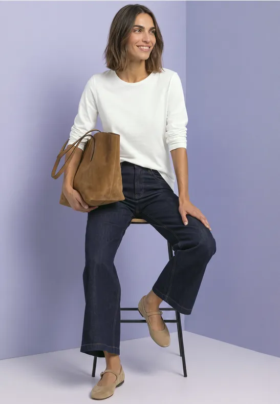 Frau in weißem Pullover und Jeans sitzt lächelnd auf einem Hocker, hält eine braune Tasche vor lila Hintergrund.