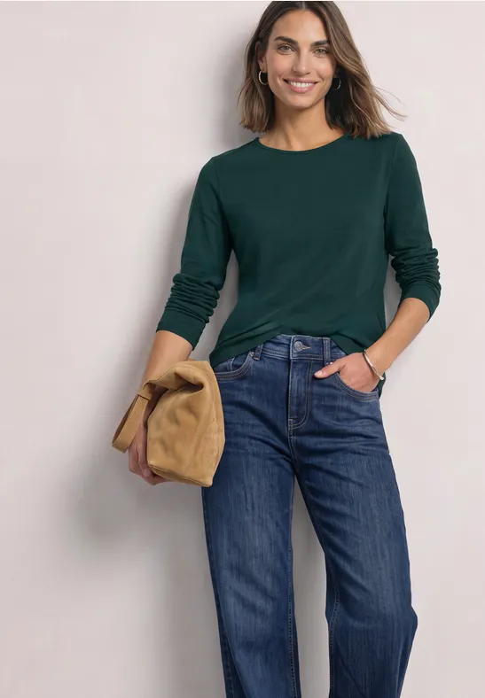 Eine lächelnde Frau in grünem Pullover, blauen Jeans und mit einer braunen Tasche