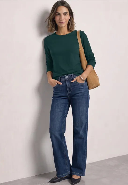Frau trägt grünen Pullover, blaue Jeans und braune Schultertasche vor hellem Hintergrund.