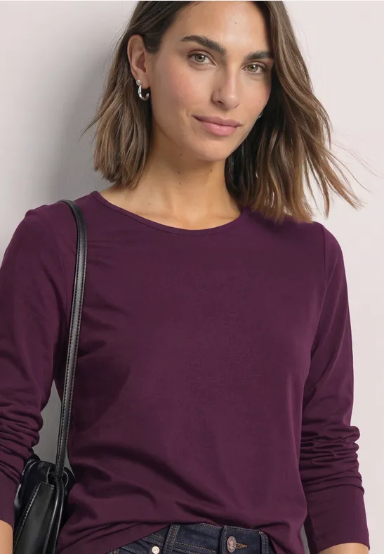 Junge Frau trägt ein bordeauxfarbenes Langarmshirt und eine schwarze Schultertasche, vor hellem Hintergrund.
