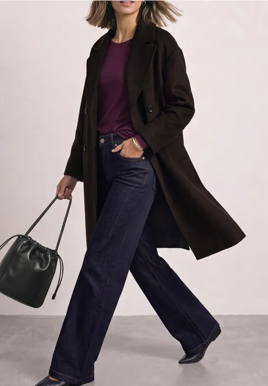 Frau in dunklem Mantel, lila Shirt, weiten Jeans und Tasche.
