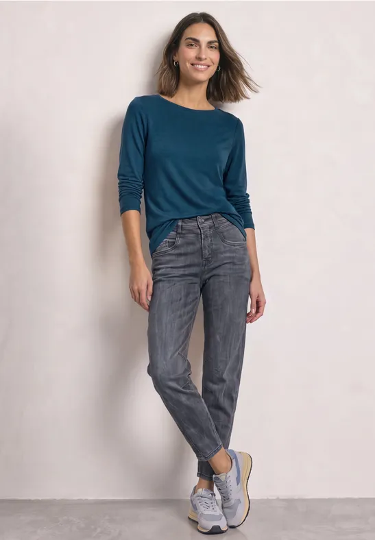 Eine Frau trägt ein blaugrünes Shirt, graue Jeans und weiße Sneakers vor hellem Hintergrund.