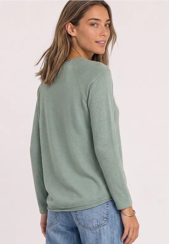 Femme en pull vert à manches longues et jean regarde par-dessus son épaule.